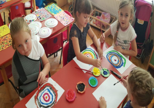 dziewczynki malują farbami obrazki z kropką a między nimi stoją w rządku małe talerzyki z farbami, w tle widać gotowe prace z kropkami